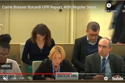 [ai] A woman speaking at a podium during a session, with a report titled 'Burundi UPR Report' in front of her. Two colleagues are seated nearby, and a person is seated in the background, focused on their device.